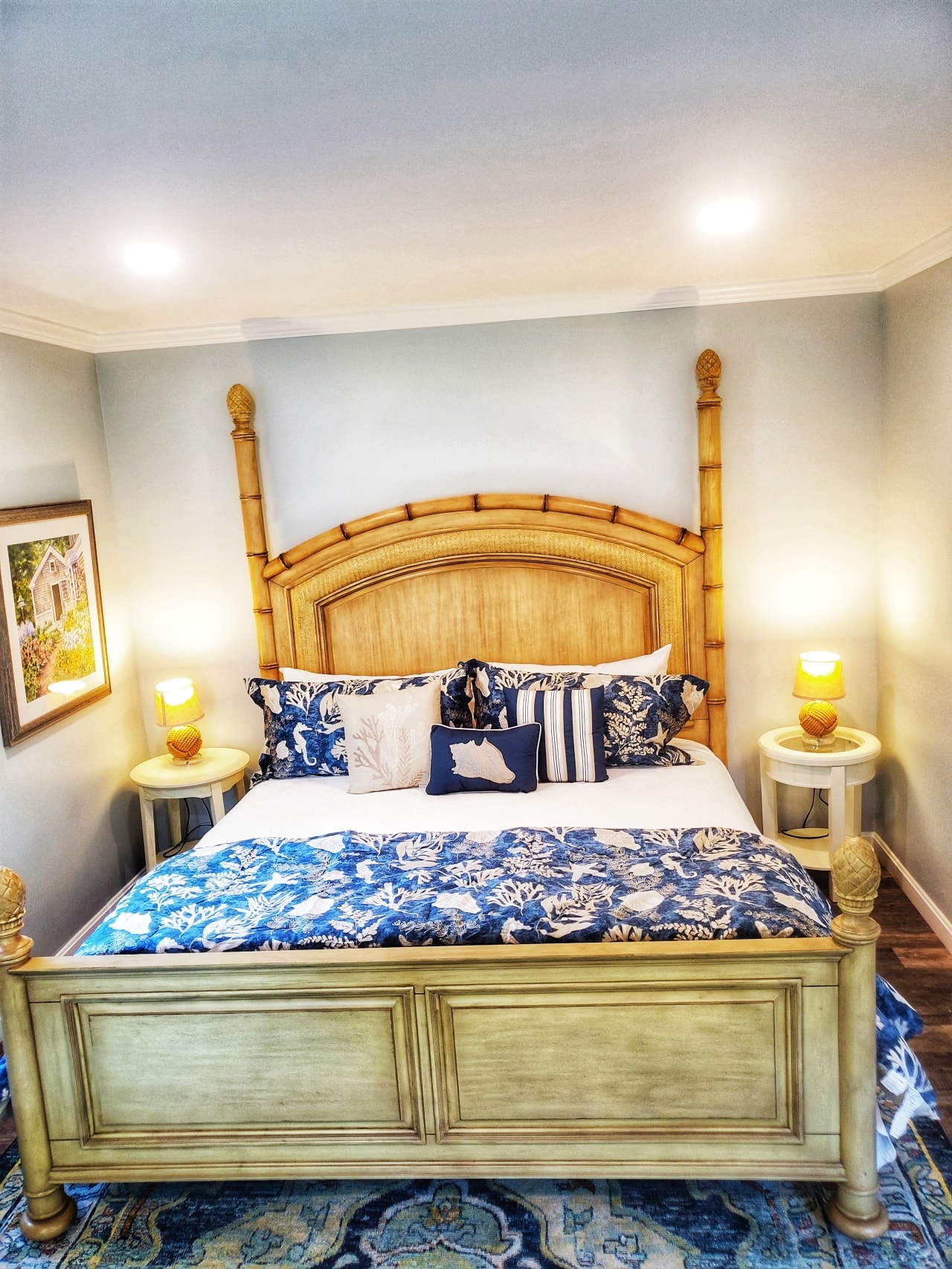 Pale beachy Tommy Bahama brand King size bed with navy blue and shells bedding and decorative pillows, round white side tables with rope wrapped lamps, blue and yellow coordinating rug, pale blue grey walls and 2 recessed lights above