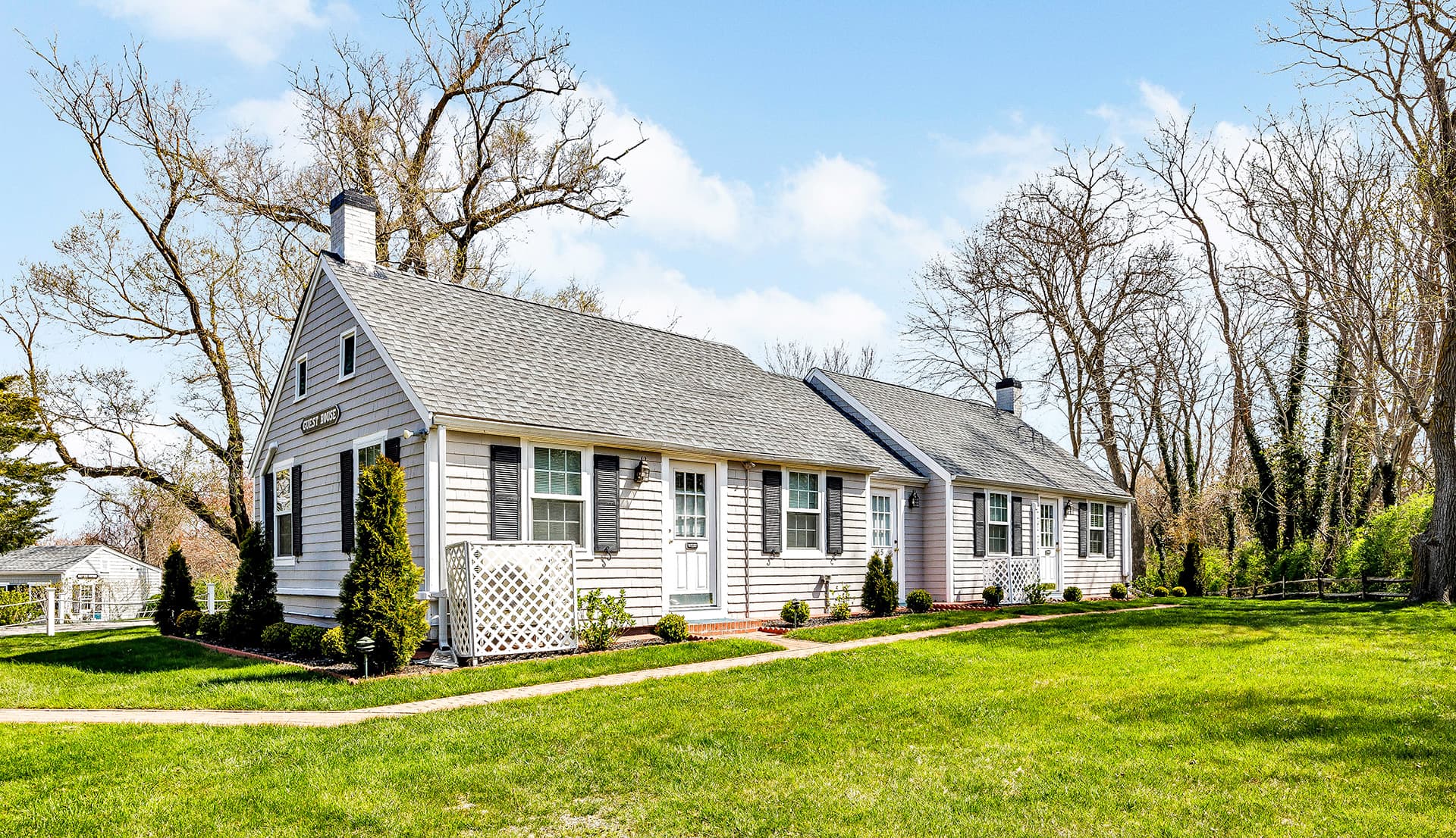 A charming guest house with a gray shingle exterior, surrounded by greenery and trees.
