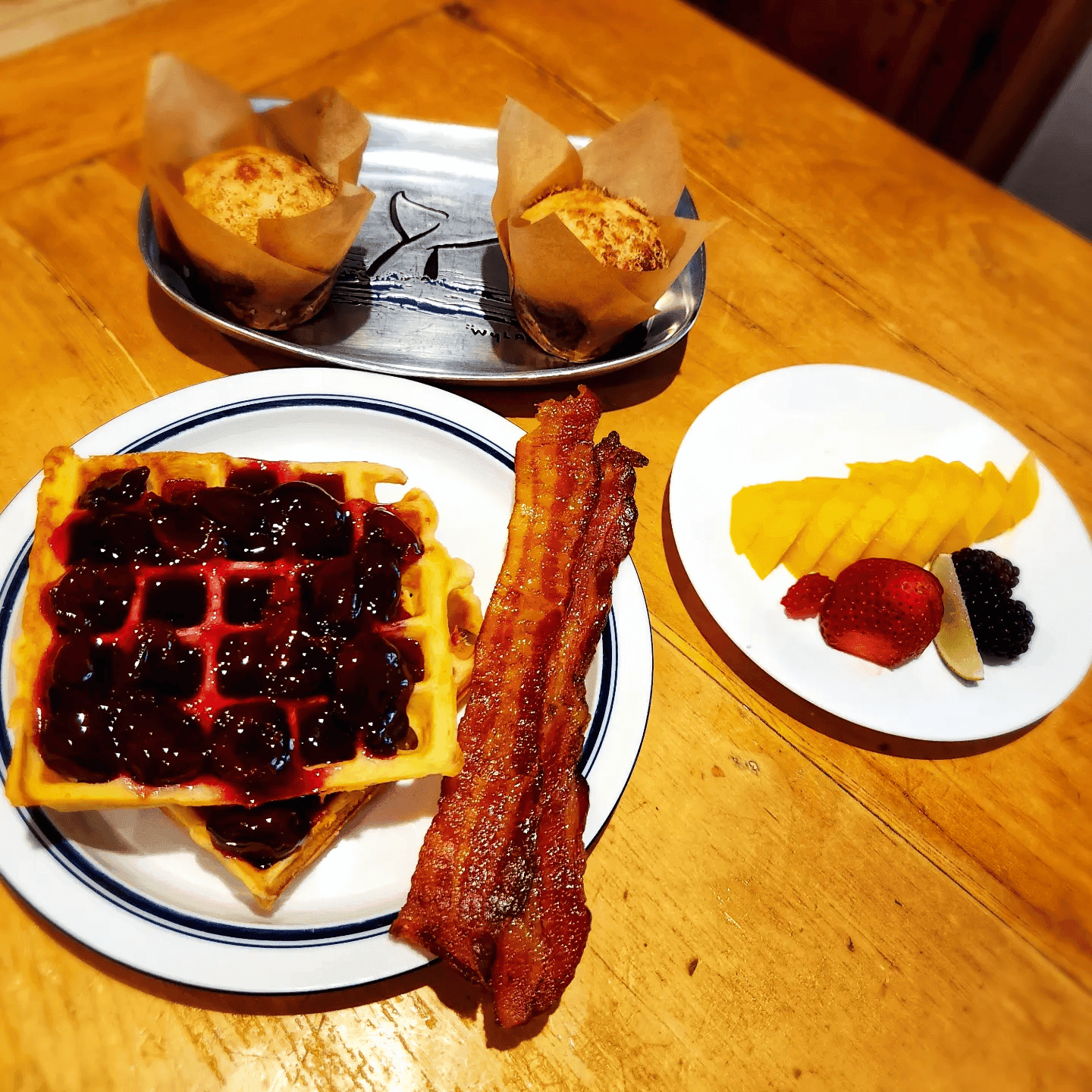 A plate of waffles topped with berry sauce, crispy bacon, accompanied by muffins and a side of assorted fresh fruit.