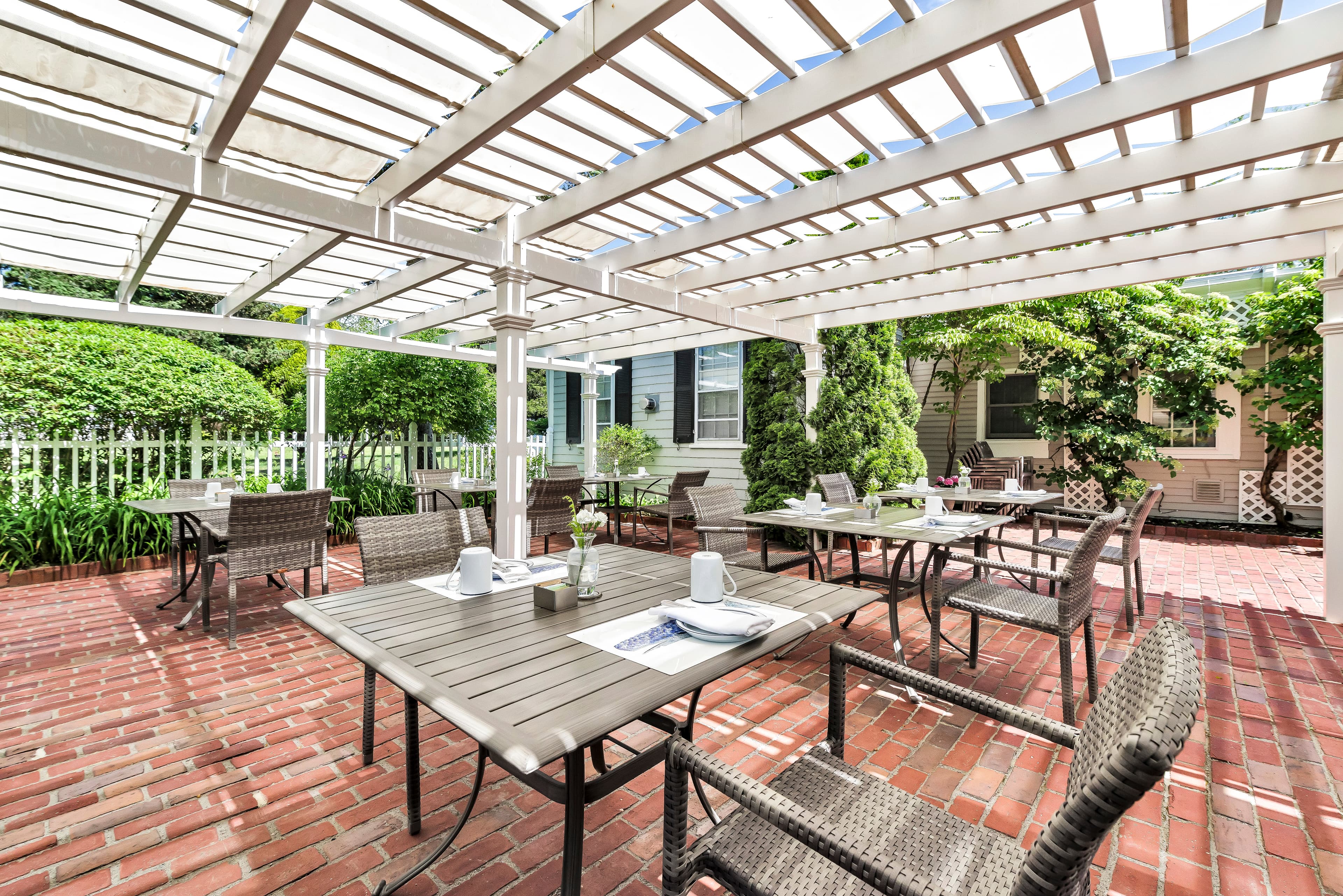 Outdoor dining area with tables and chairs under a pergola, surrounded by greenery.