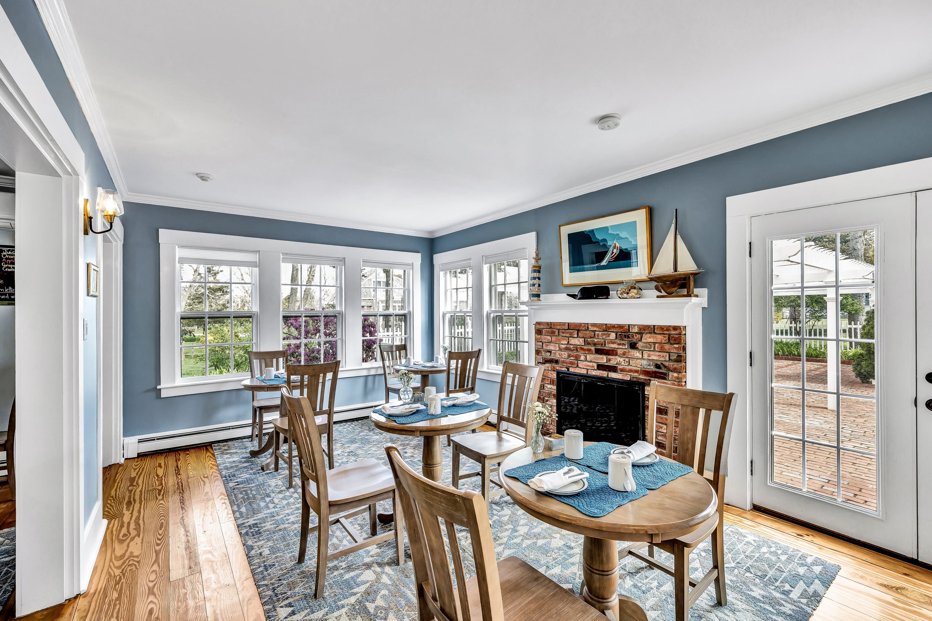 A bright, inviting dining room featuring wooden tables and chairs, a brick fireplace, and large windows letting in natural light.