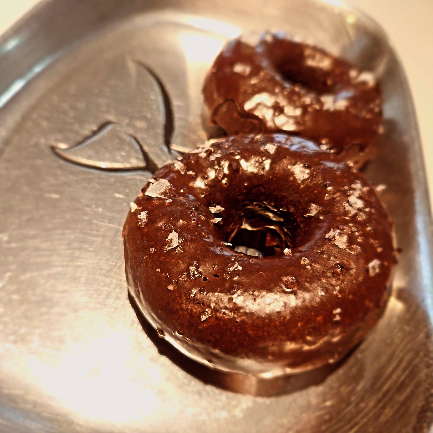 Two chocolate donuts topped with sea salt on a silver tray.