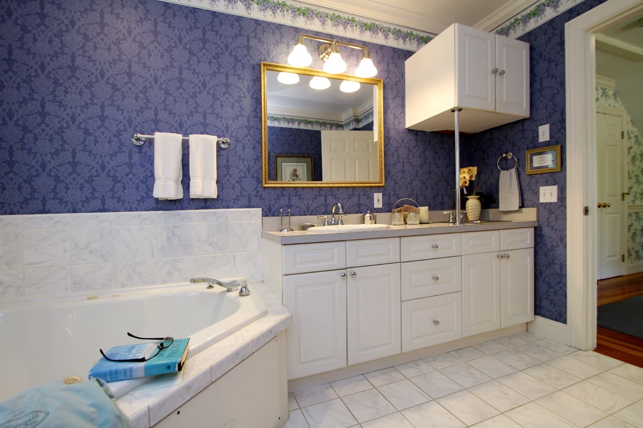 another view of the bathroom with tub, vanity with sink, mirror above, and refrigerator tucked away in cabinet, with towel bar and towels and purple brocade wallpaper