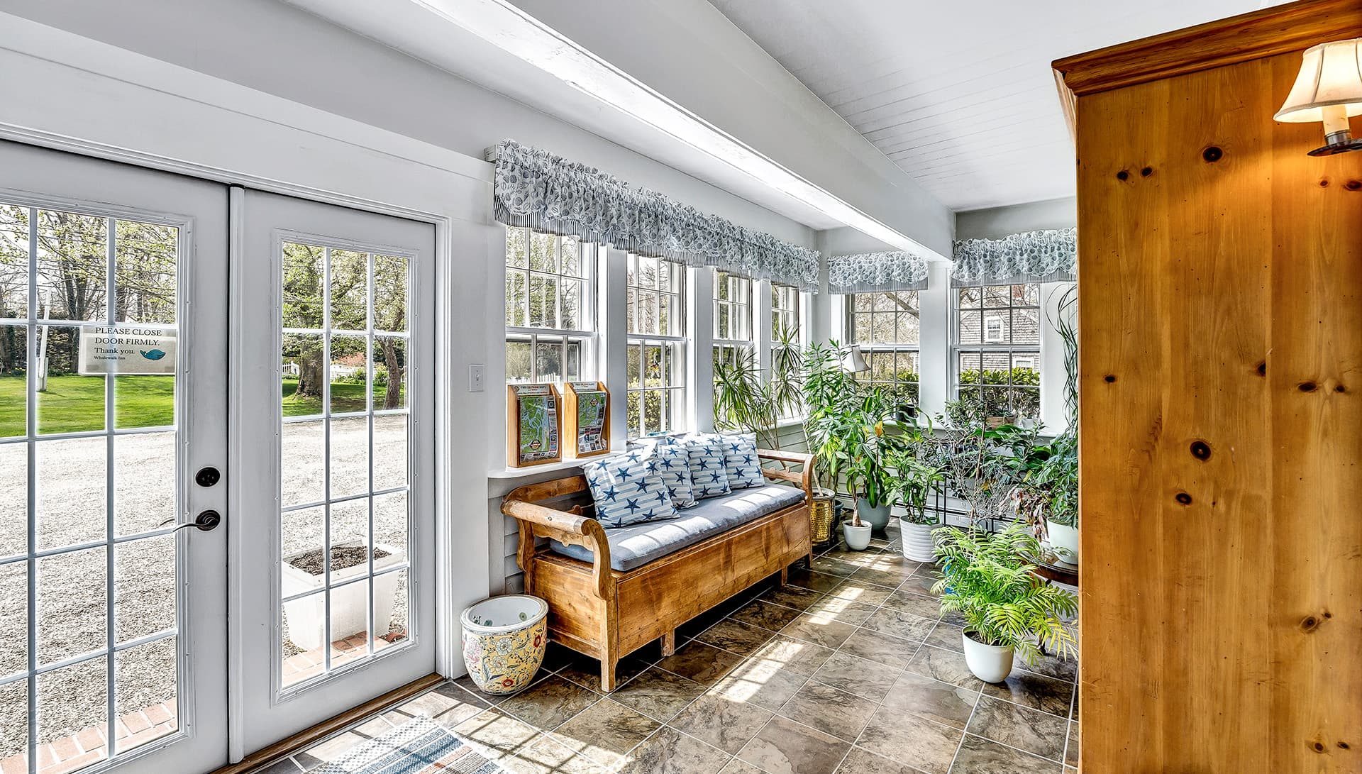 A bright sunroom with a wooden bench, potted plants, and double doors leading outside.
