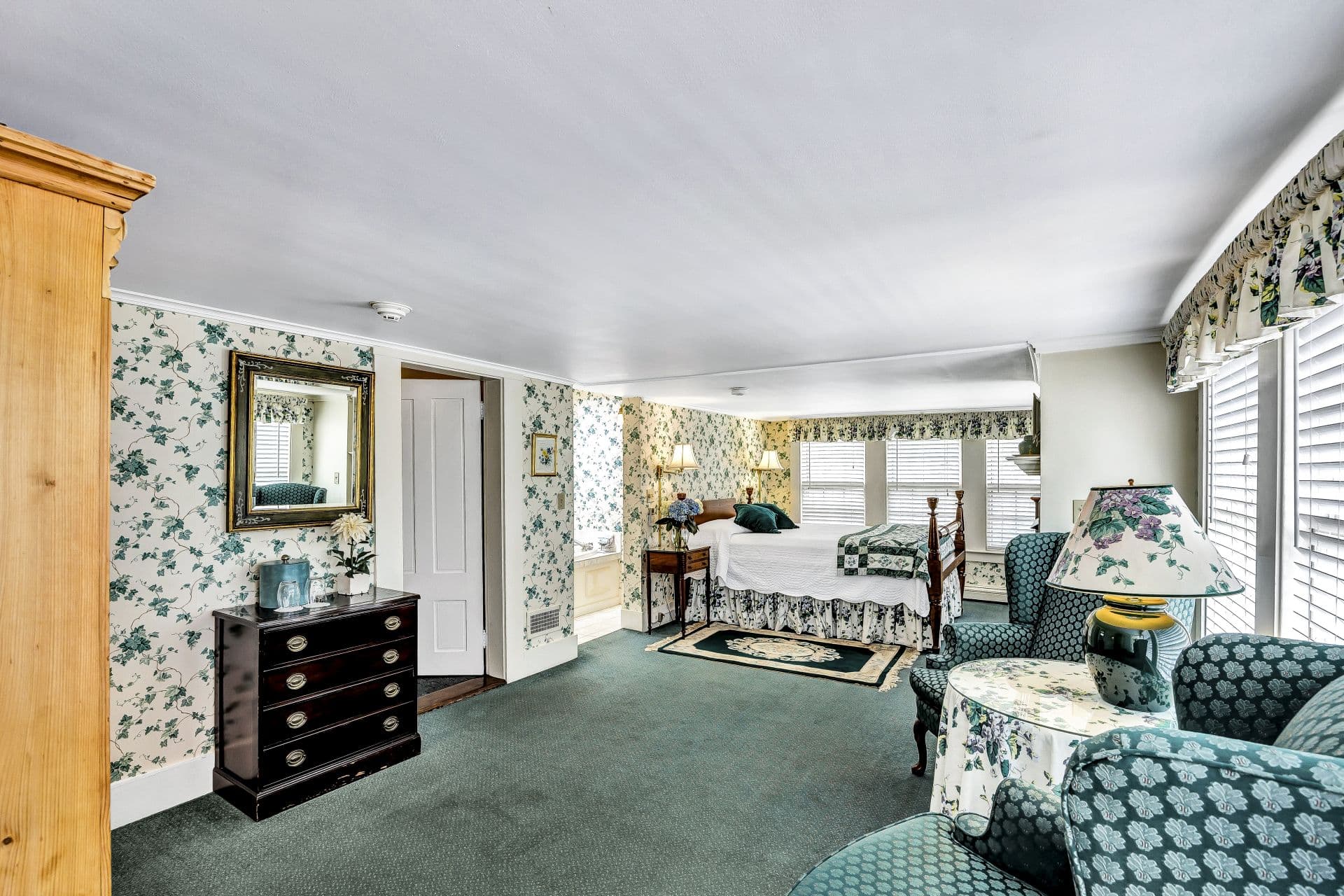 A spacious bedroom with blue-green carpet and floral wallpaper. The room has a bed, a dark wood dresser, and a seating area with two chairs.