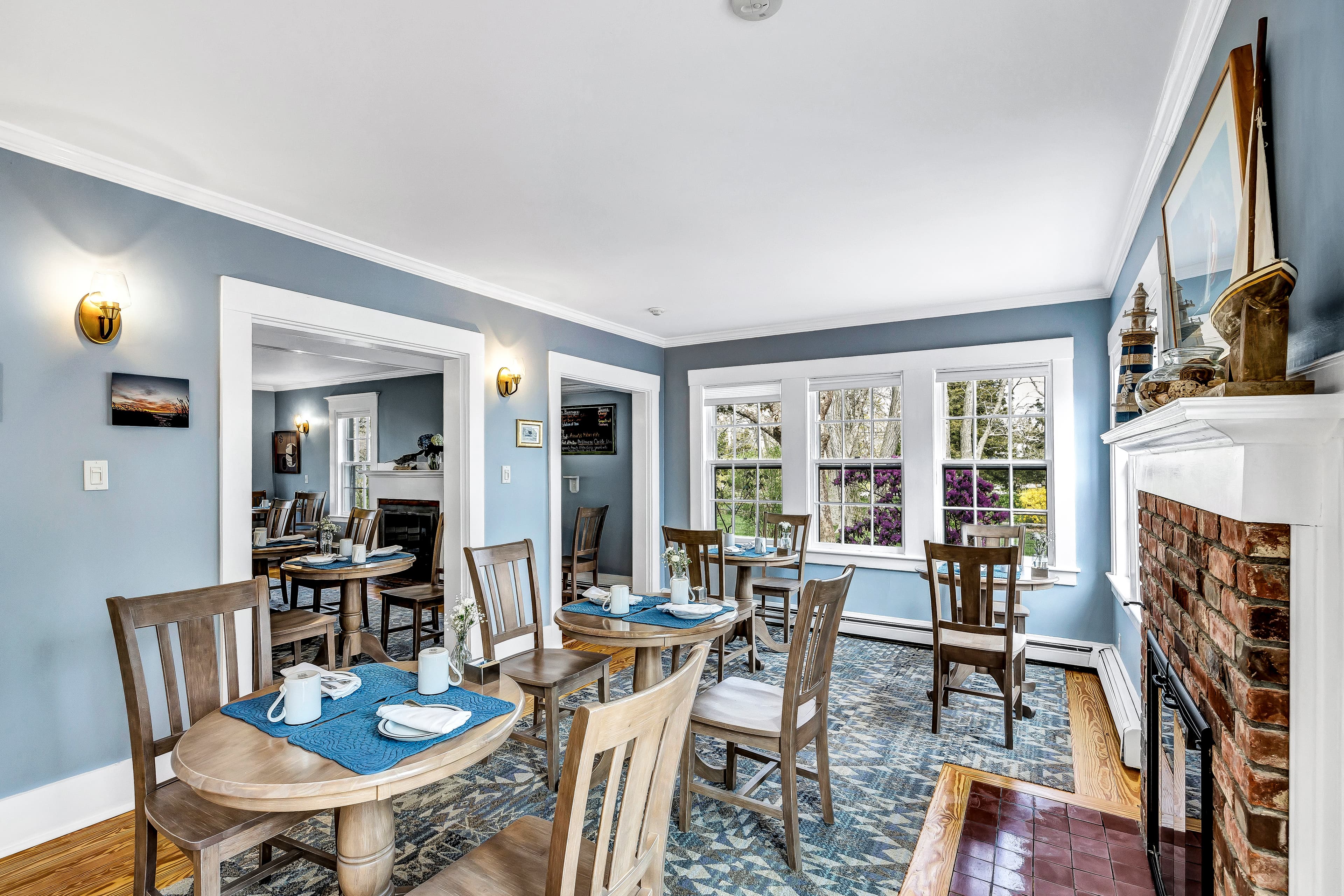 Cozy dining area with wooden tables and a blue accent wall, featuring large windows and a brick fireplace.