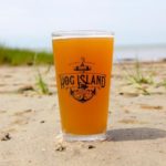 A glass with the Hog Island logo filled with a light golden beer and sitting in the sand.