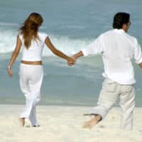 Couple wearing white shirts and pants walking long the beach as waves roll in.